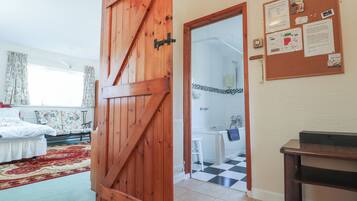 Una habitación con una puerta de madera que da a un baño con piso a cuadros. Hay un tablón de anuncios en la pared con varios avisos y una pequeña mesa con una silla.