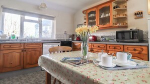A kitchen with wooden cabinets, a microwave, and a dining table with a floral tablecloth. - Otter Cottage (Newport)