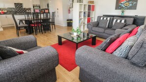 Una sala de estar moderna con sofás grises, una mesa de centro de vidrio y una alfombra roja. Hay un comedor con una mesa y sillas negras, y una cocina al fondo.