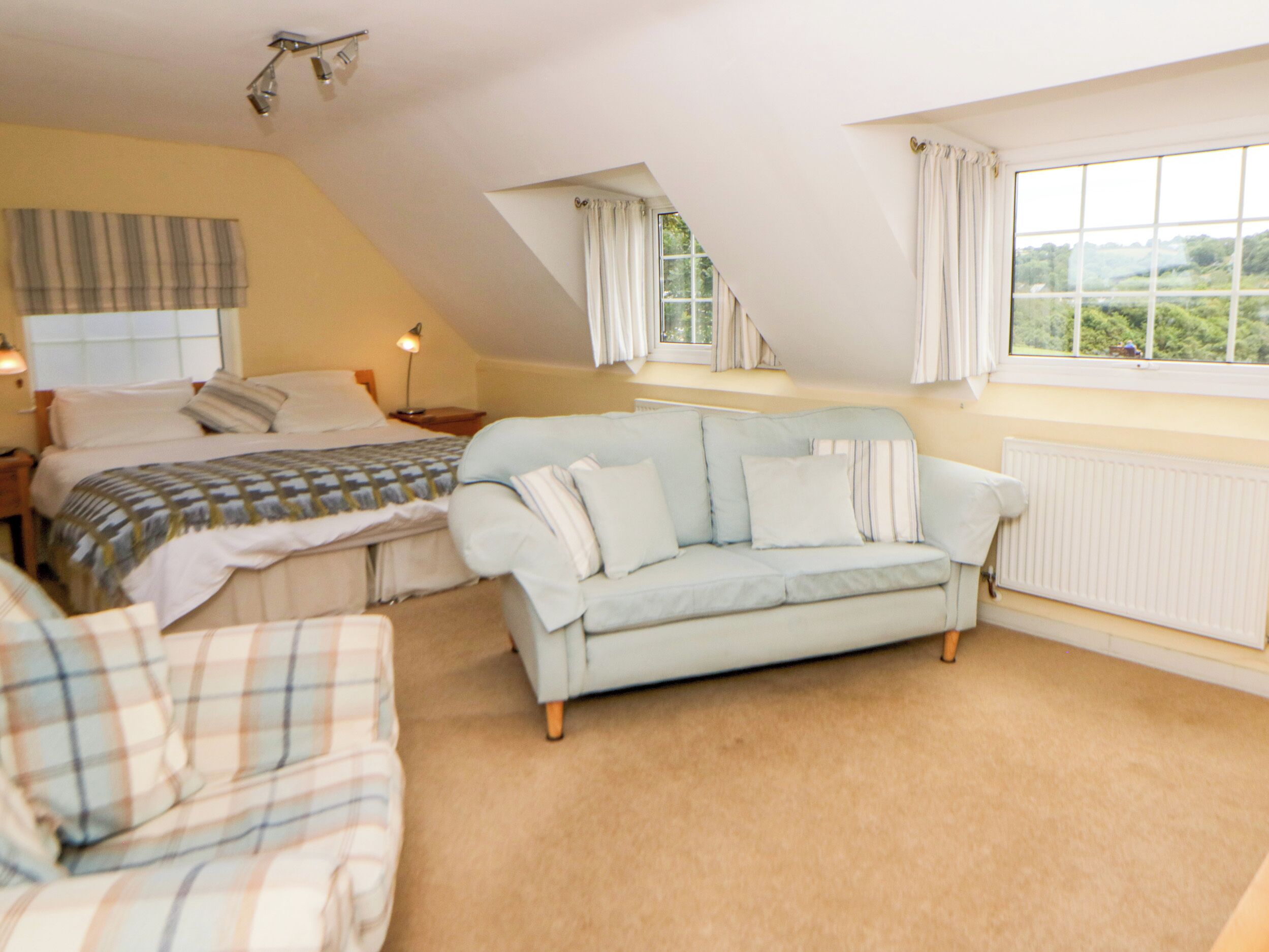 A bedroom with a bed, a light blue sofa, a plaid chair, a nightstand, a lamp, and a window with curtains.