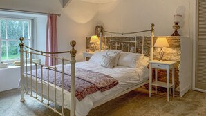 A bedroom with a white four-poster bed, two bedside tables with lamps, a stone wall, a window with a pink curtain, and a radiator. - The Farmhouse (Cardigan)