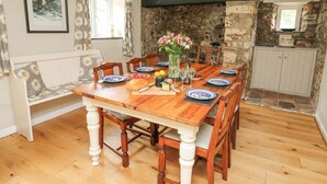 A dining room with a wooden table set for six, surrounded by chairs. A stone wall and a window with curtains are visible in the background. - The Farmhouse (Cardigan)