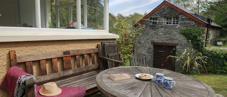 A wooden bench with a woven seat cushion, a round wooden table with a plate of biscuits and a mug, and a stone building in the background.