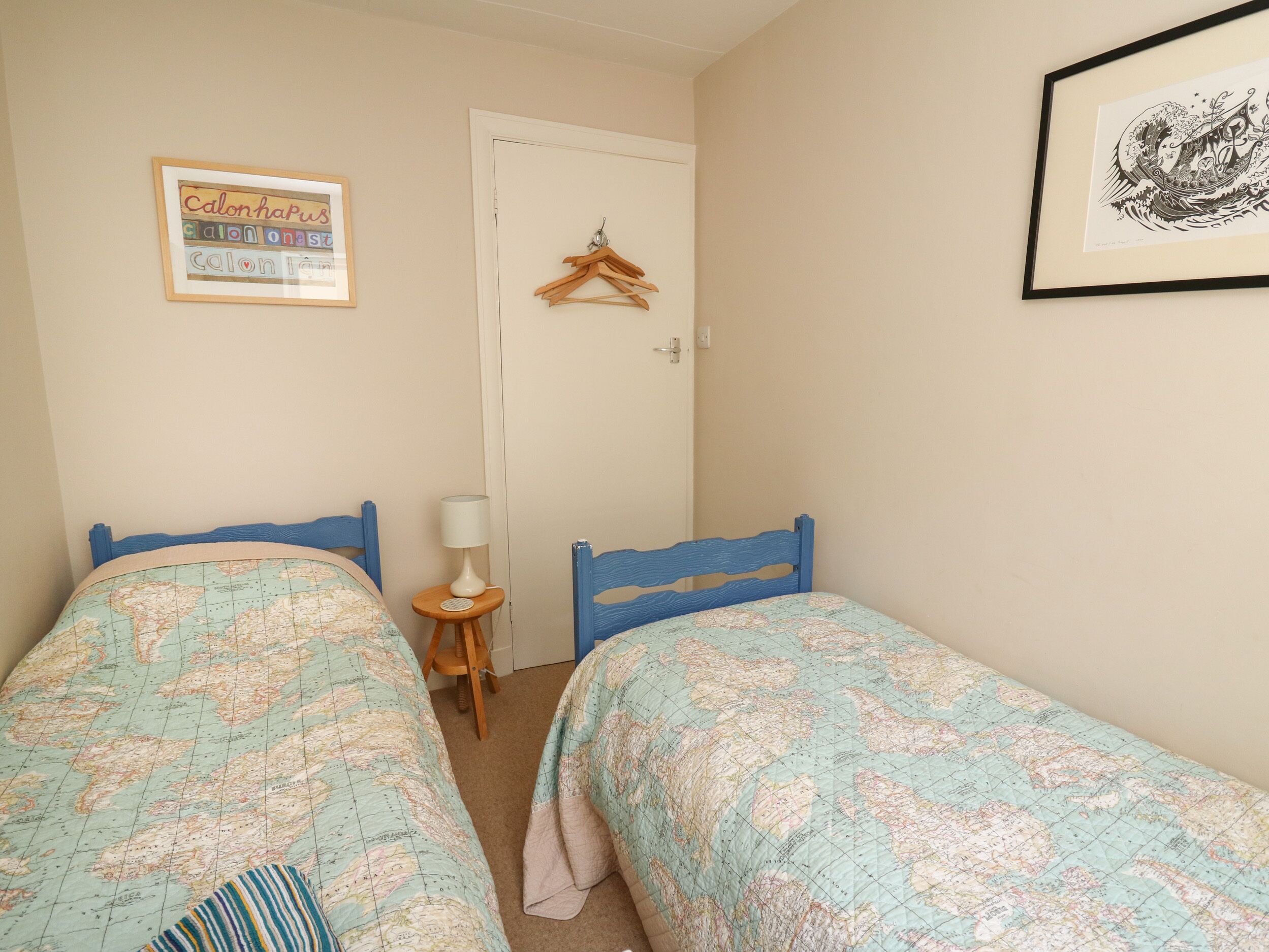 A bedroom with two beds, a small table with a lamp, a door with a hanger, and a framed picture on the wall.