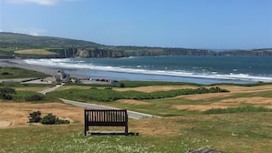 A coastal landscape with a bench overlooking the sea, a road leading to a building, and cliffs in the background.