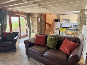 A living room with a brown leather sofa, a dining table with yellow flowers, a kitchen with stone walls, and a staircase. - Stable Cottage (Cardigan)
