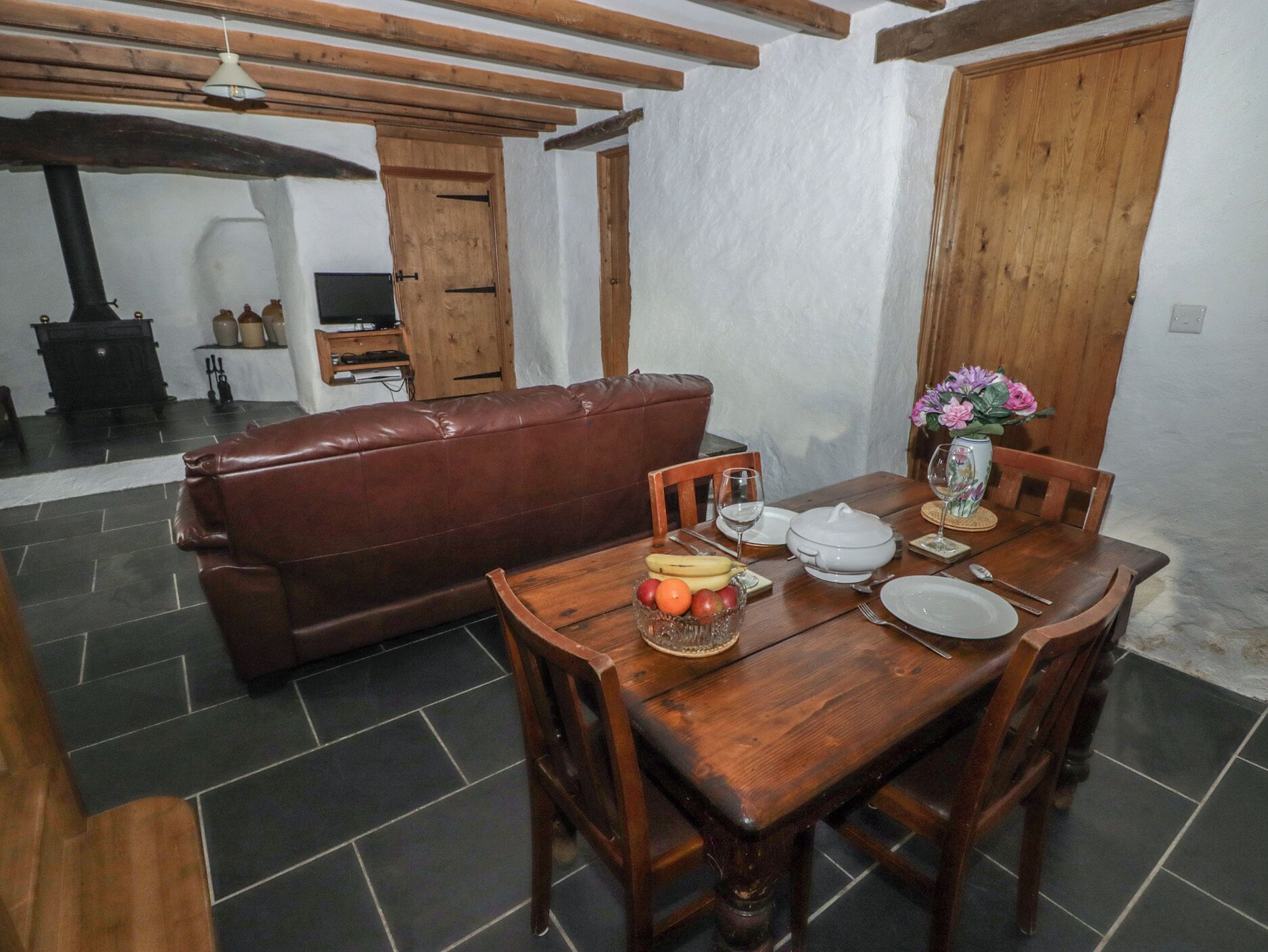 A dining area with a wooden table set for two, a brown leather sofa, and a kitchen in the background.