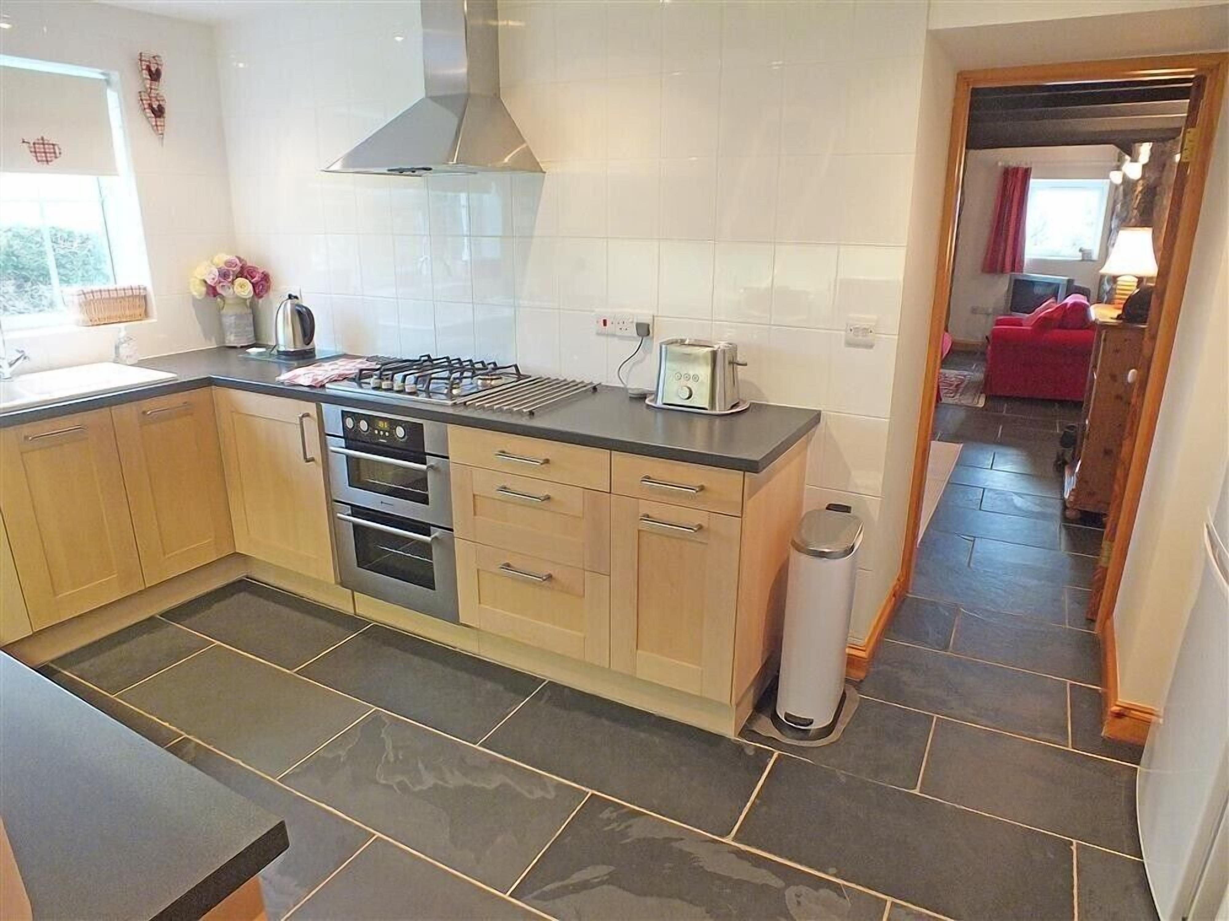 A modern kitchen with a gas stove, oven, and a toaster. There is a trash bin and a window with flowers.