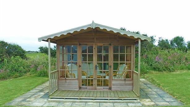 Ein hölzerner Pavillon mit Glaseinfassung, ausgestattet mit einem Tisch und Stühlen, auf einer pflasterten Terrasse in einer grünen Umgebung.