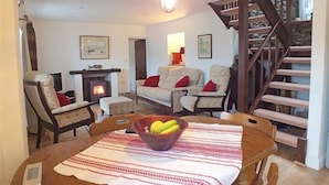 Ein gemütliches Wohnzimmer mit Kamin, bequemen Sitzmöglichkeiten, einem Esstisch mit Obstschale und einer Holztreppe.