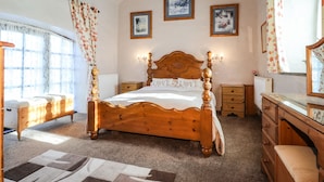 Ein Schlafzimmer mit einem hölzernen Bett, einem Nachttisch, einer Kommode und einem Spiegel. An der Wand hängen zwei gerahmte Bilder und ein Fenster mit Vorhängen.