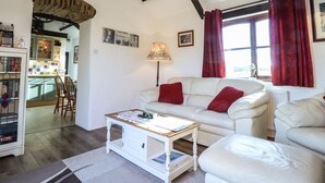 Ein Wohnzimmer mit einem weißen Sofa, roten Kissen, einem Holztisch und einer Lampe. Im Hintergrund ist eine Küche mit grünen Schränken und einer gefliesten Rückwand zu sehen.