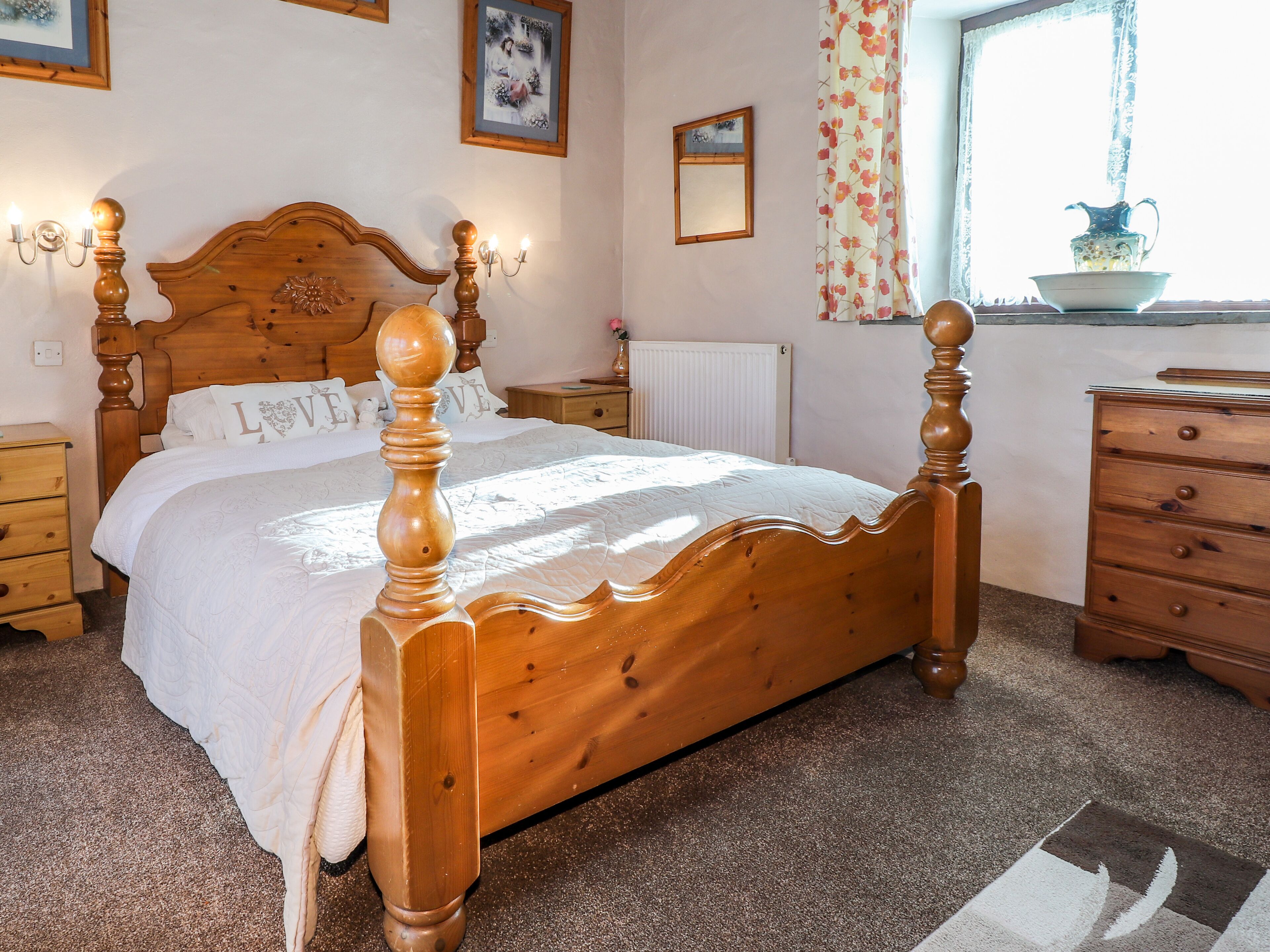 A wooden four-poster bed with a white bedspread, a nightstand with a lamp, a dresser, and a window with a view of the outdoors.