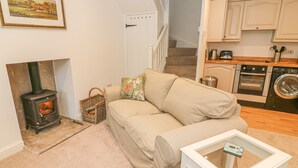 A cozy living room with a sofa, a glass coffee table, a fireplace, and a kitchen area with appliances. - The Little Bower (Hawes)