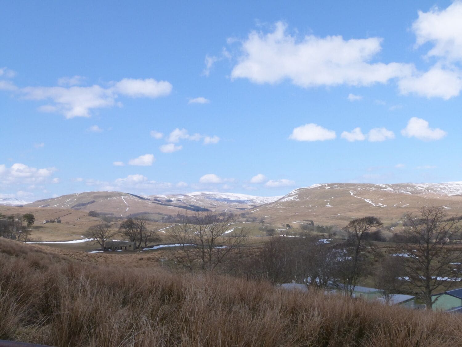 A rural landscape with rolling hills, sparse vegetation, and a few buildings in the distance.