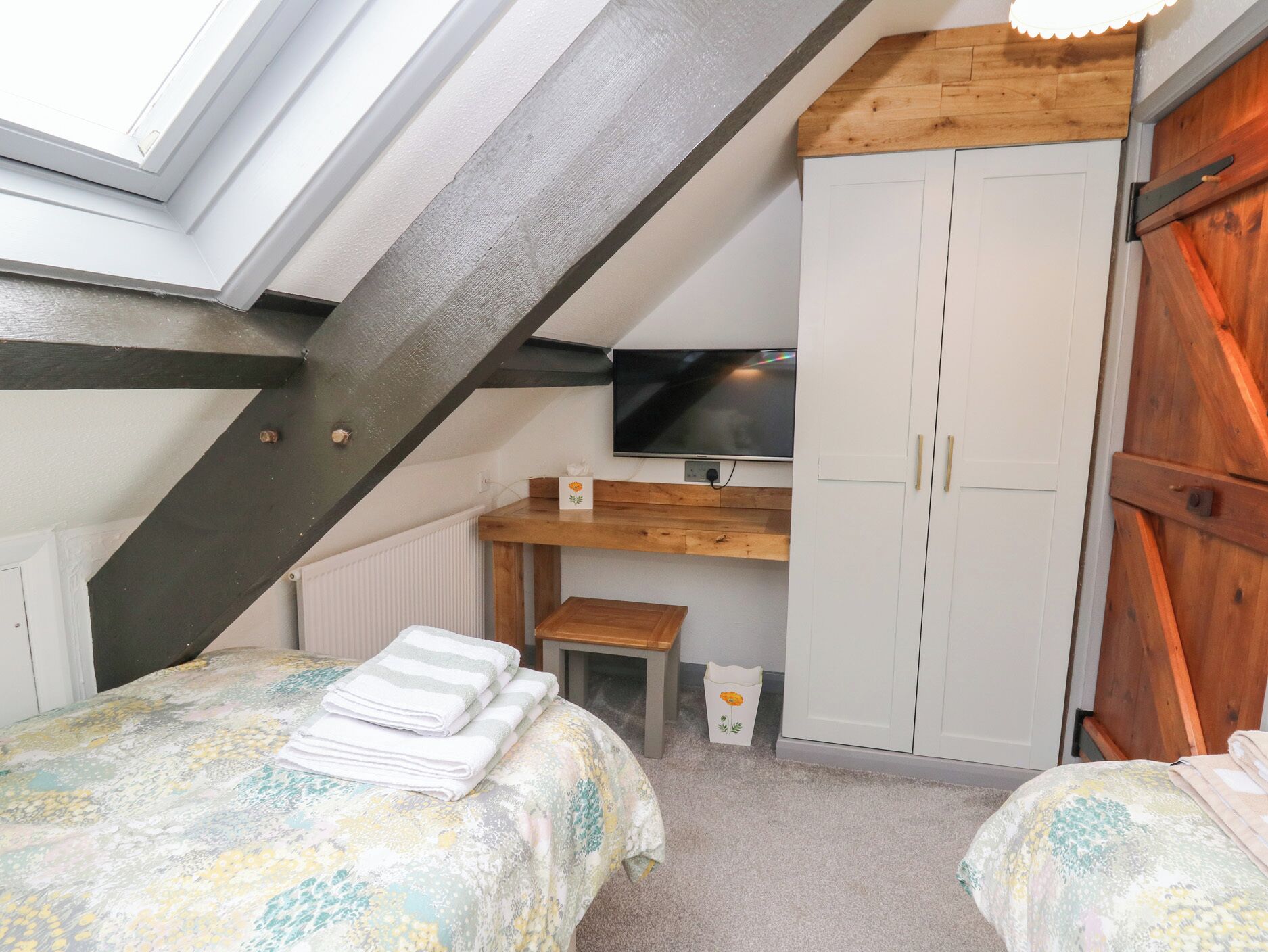 A bedroom with a sloped ceiling, a bed with a floral bedspread, a wooden desk, a television, and a wardrobe.