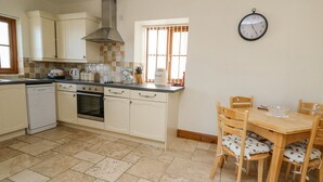 A kitchen with white cabinets, a stove, oven, and a dining area with wooden chairs and a table. - Beudy (Cardigan)
