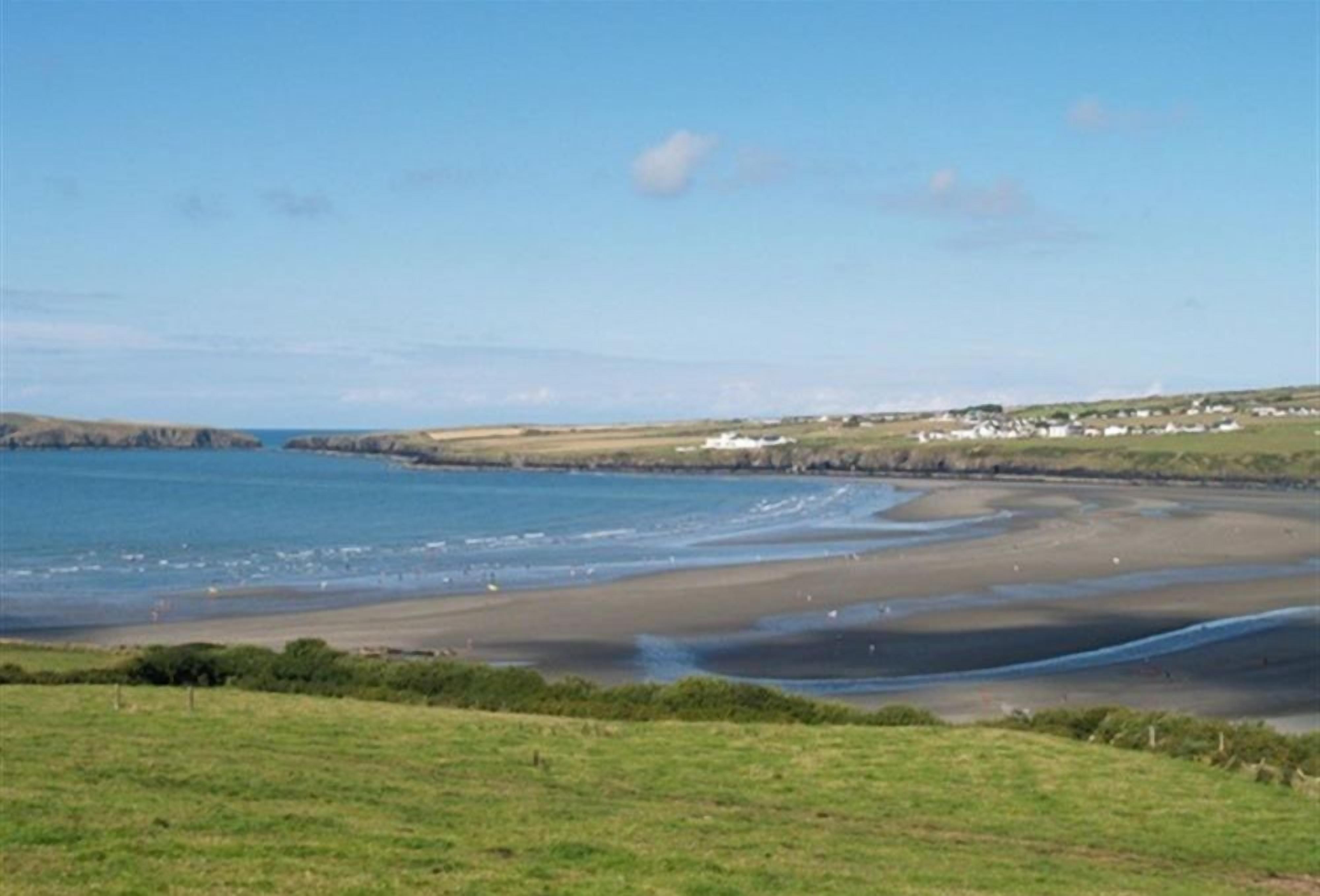 A coastal landscape with a sandy beach, grassy fields, and a distant view of houses along the shore.