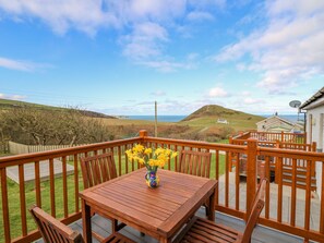 A wooden deck with a table and chairs, a vase of yellow flowers, and a scenic view of hills and the sea. - Llaethdy (Cardigan)