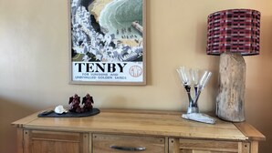 A framed poster of Tenby, a wooden cabinet with drawers, a decorative lamp, and a small vase with glass objects on a table.