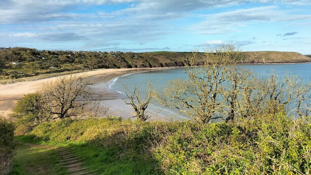 A coastal landscape with a sandy beach, a calm sea, and a hilly background.