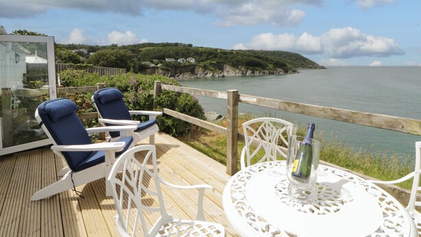 Een terras met twee blauwe stoelen en een witte tafel met een fles. Een houten hek en een bord zijn zichtbaar.