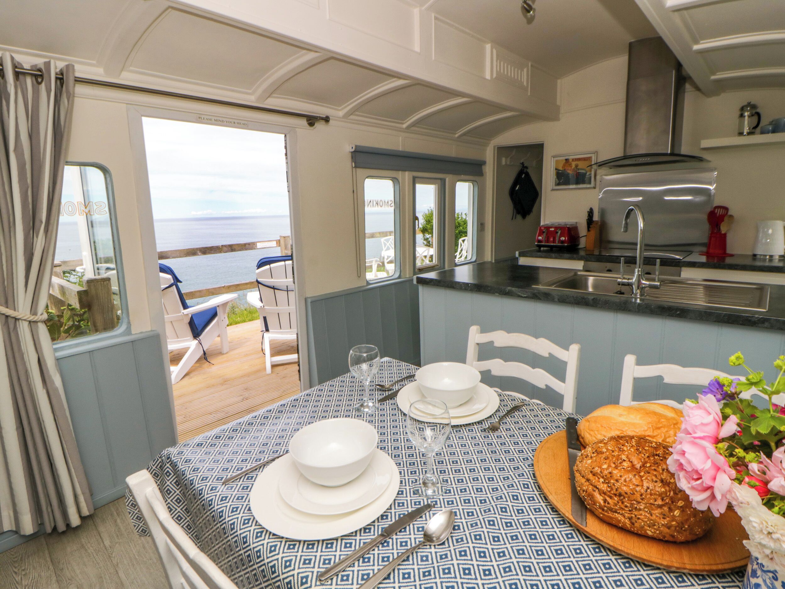 A dining area with a table set for two, a kitchen with a toaster, and a view of the sea through the open door.