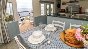Een eetgedeelte met een tafel gedekt voor twee personen, een keuken met een broodrooster, en uitzicht op zee door de open deur.