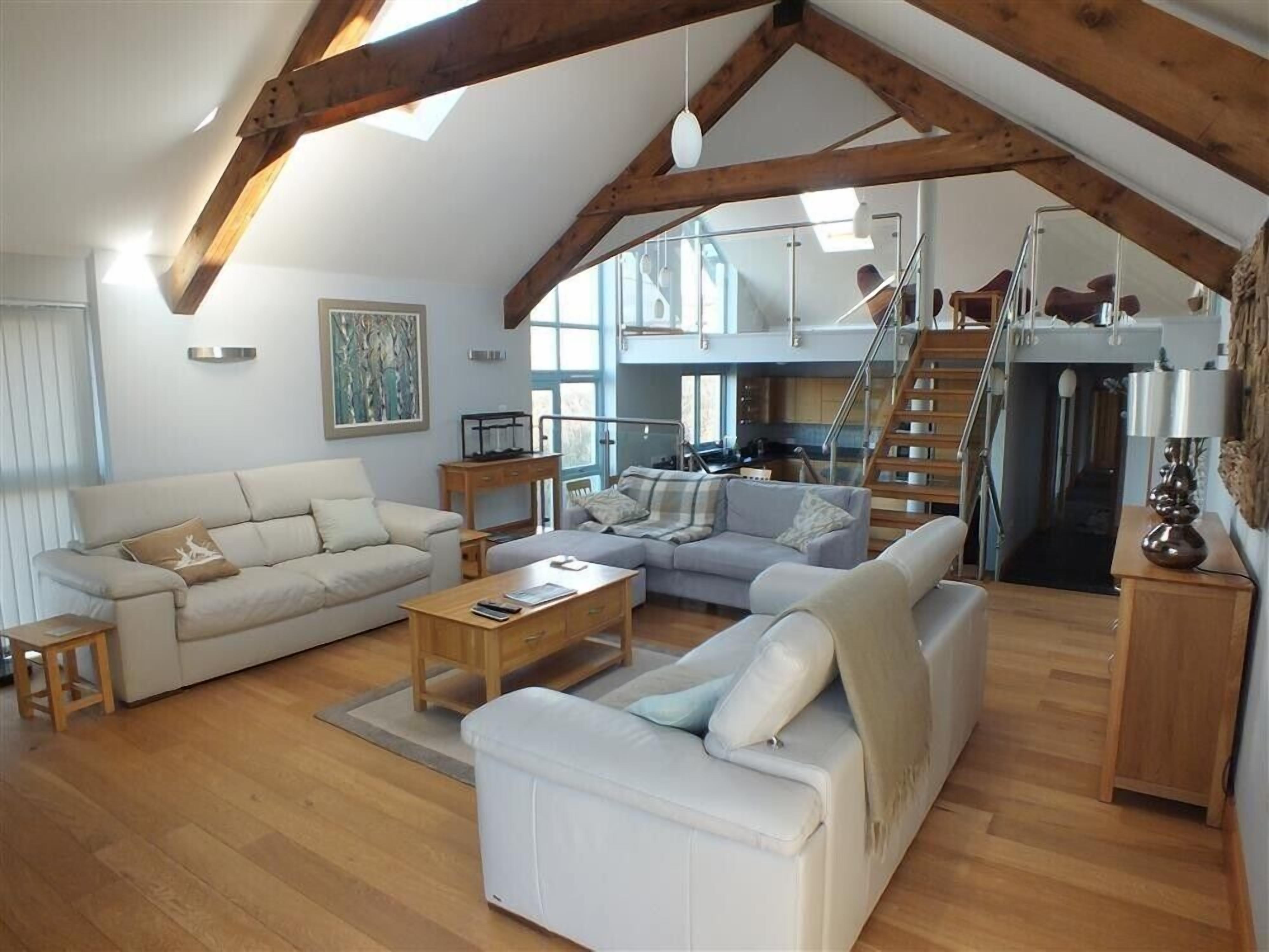 A spacious loft with high ceilings, exposed wooden beams, a living area with sofas and a coffee table, a kitchen with a staircase, and a large window providing natural light.