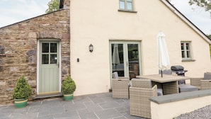 Un bâtiment doté d’une façade en pierre, d’une porte verte et d’un patio aménagé avec des meubles en osier et un barbecue.