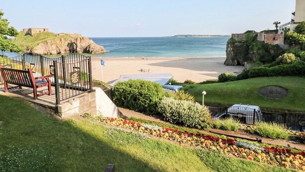 Une vue sur la côte, avec une plage, un jardin aux fleurs multicolores et une construction en pierre en arrière-plan.