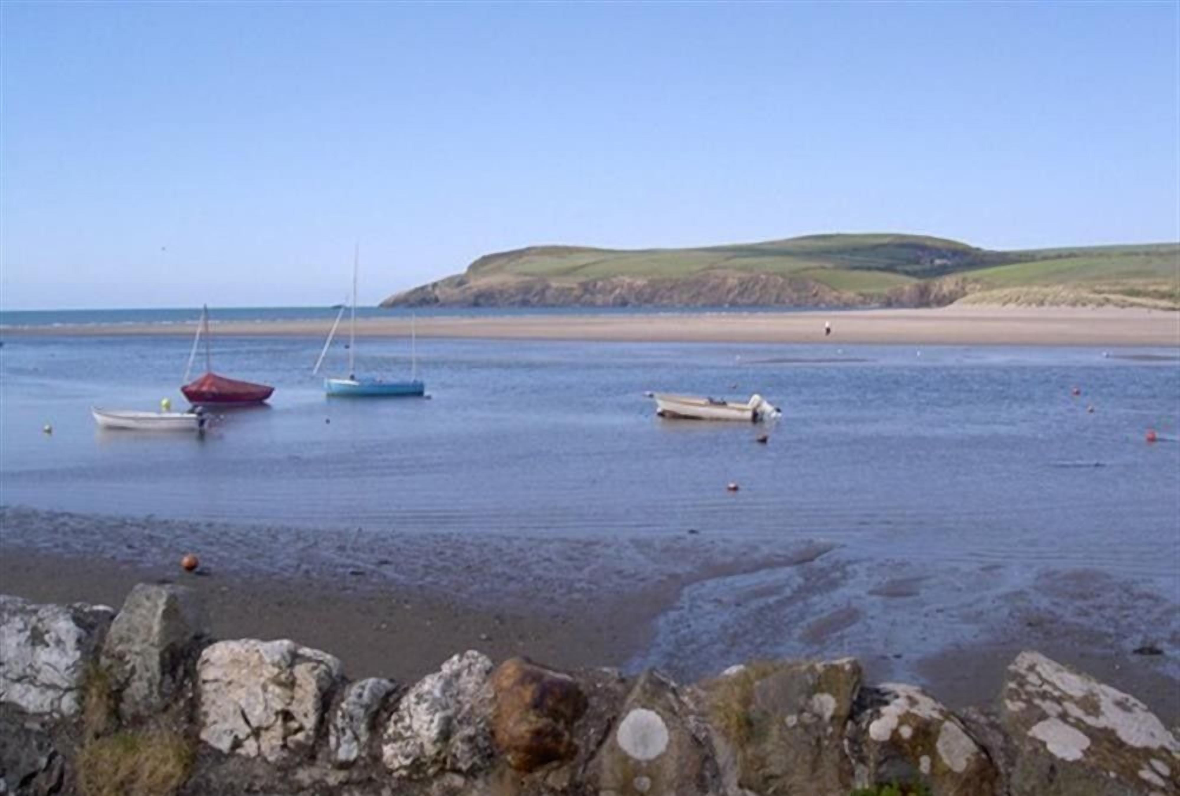 A coastal scene with boats on calm water, a sandy beach, and a stone wall in the foreground.