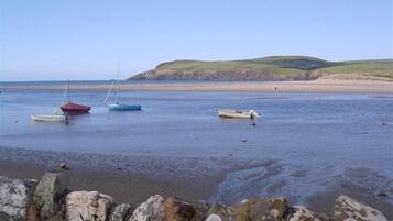 A coastal scene with boats on calm water, a sandy beach, and a stone wall in the foreground.