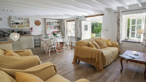 Uma sala de estar acolhedora com um sofá, uma mesa de jantar com cadeiras, uma área de cozinha com micro-ondas e estantes, e uma janela grande com cortinas.