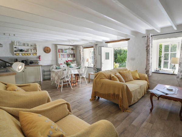 A cozy living room with a sofa, a dining table with chairs, a kitchen area with a microwave and bookshelves, and a large window with curtains. - End Cottage (Newport)