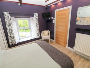 Quarto com cama, janela com cortinas, uma cadeira, porta de madeira, televisão e radiador.