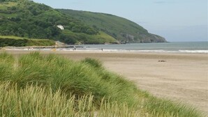 A beach with a grassy foreground, a sandy beach, and a hill with a building in the background. - Gellifor (Cardigan)