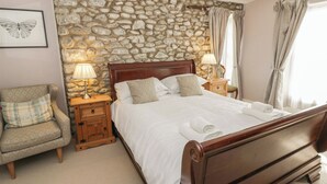 A bedroom with a stone wall, a wooden bed, two bedside tables with lamps, a chair, and a window with curtains. - Dewi Villa (Aberaeron)