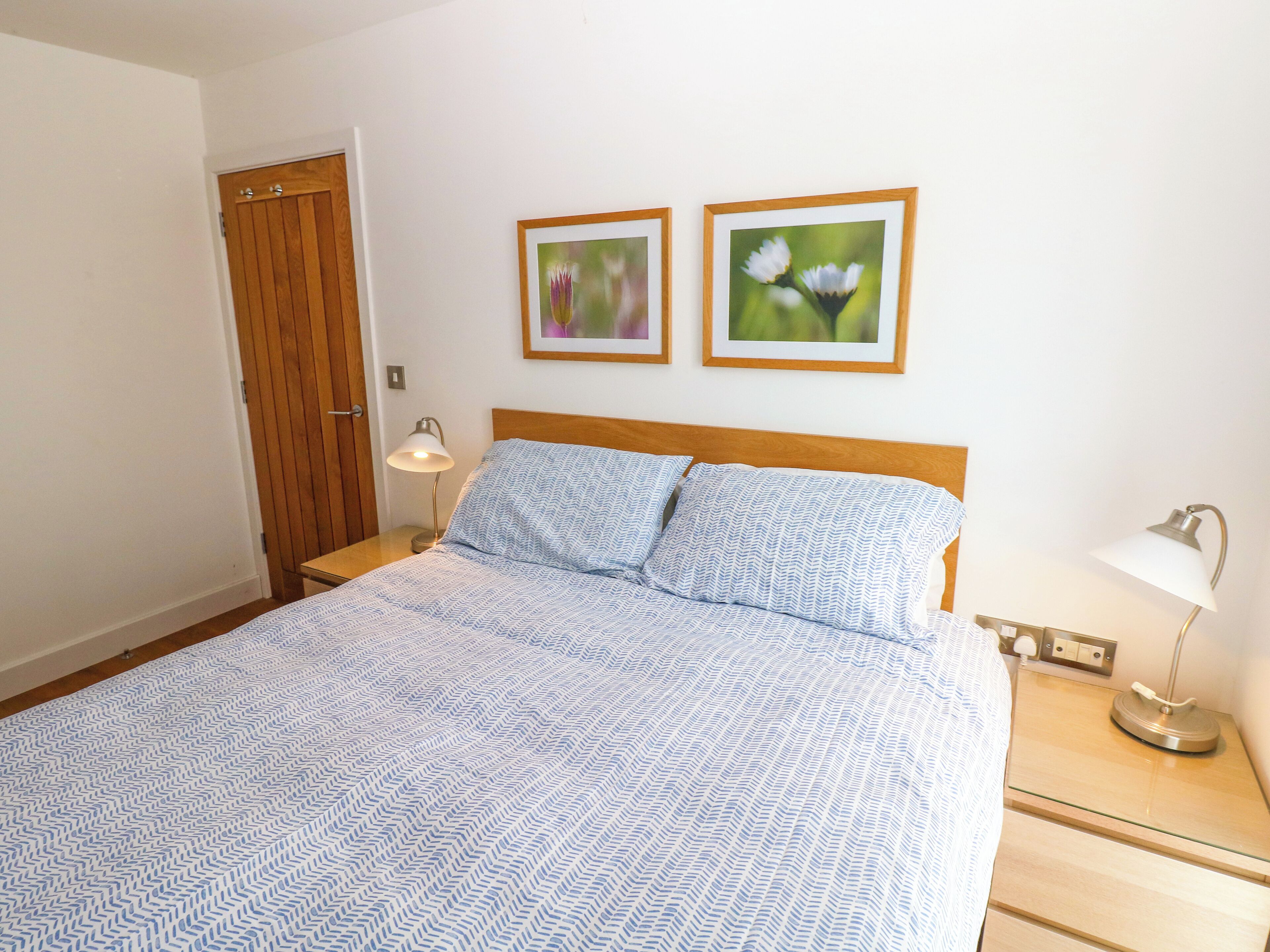 A bedroom with a bed, two bedside lamps, a wooden door, and two framed pictures on the wall.