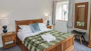 Cama bem arrumada com colcha verde estampada, duas luminárias de cabeceira, mesa de cabeceira de madeira, uma cadeira, uma janela com cortinas e um espelho na parede.
