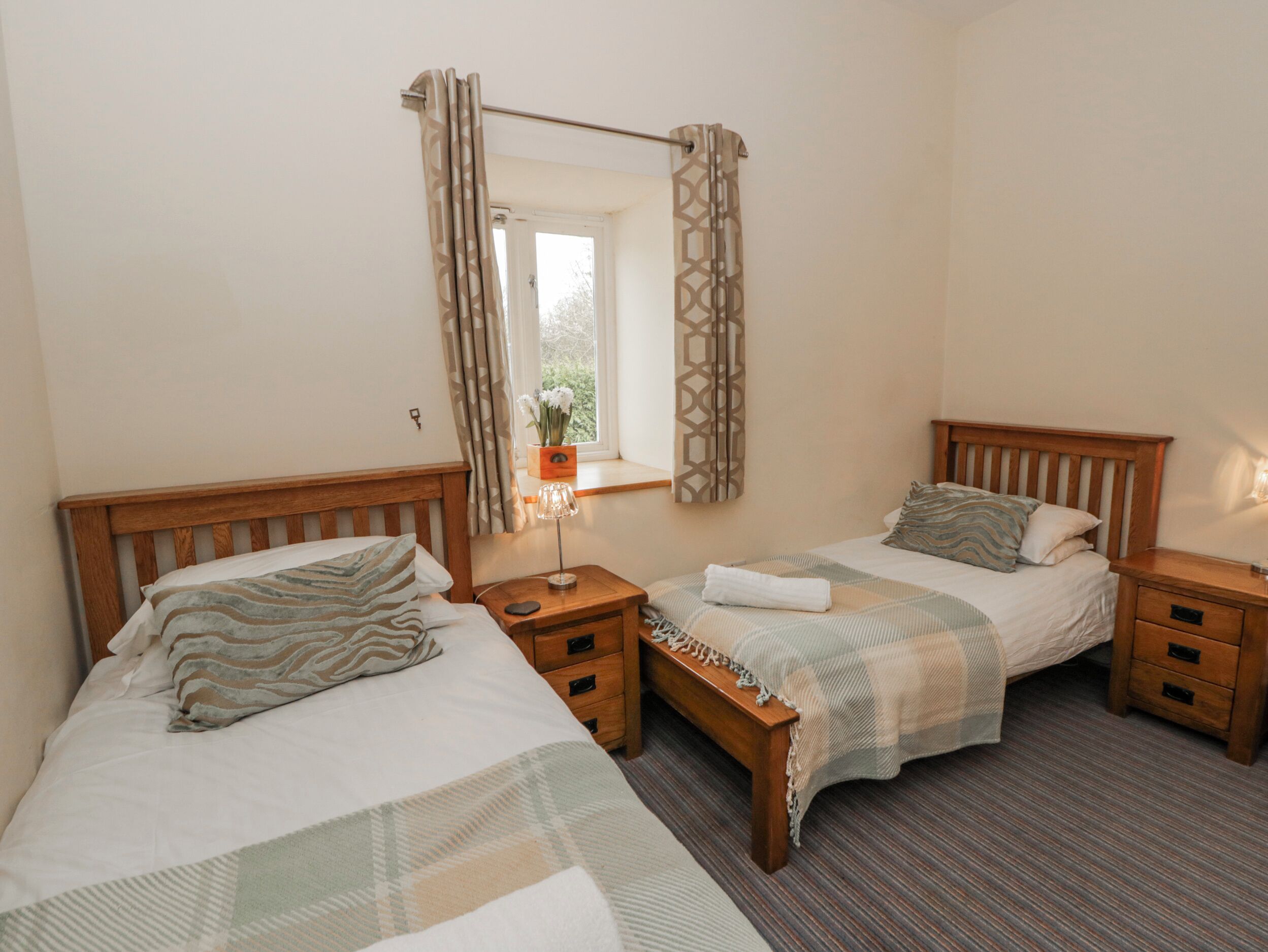 A bedroom with two single beds, wooden headboards, bedside tables with drawers, a bedside lamp, and a window with curtains.