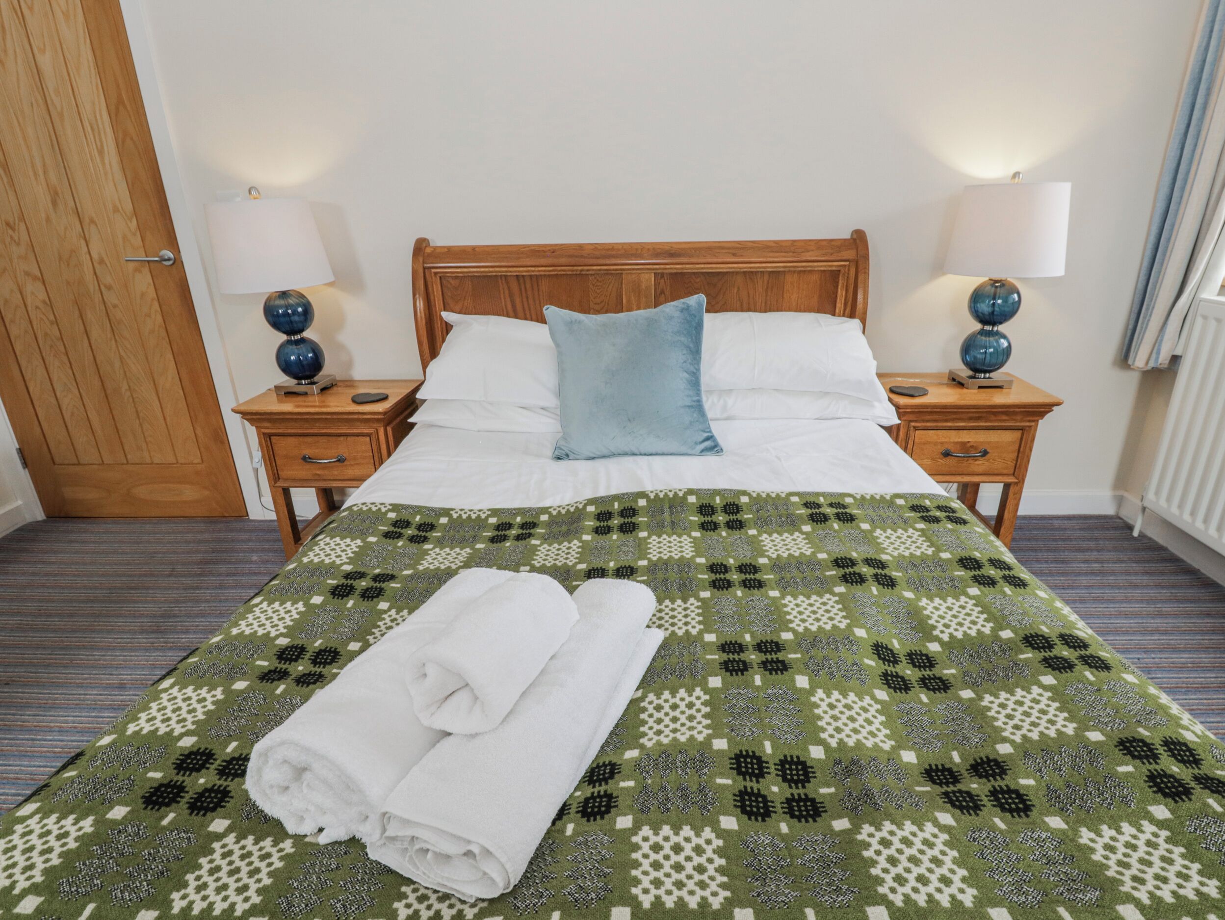 A neatly made bed with a patterned bedspread, two pillows, and a folded towel on a wooden nightstand.