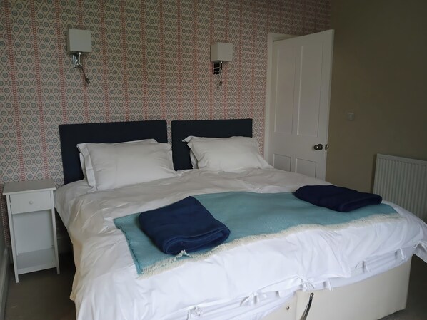 A neatly made bed with white linens and two blue towels, a white bedside table, a wall-mounted light, and a door to the right.