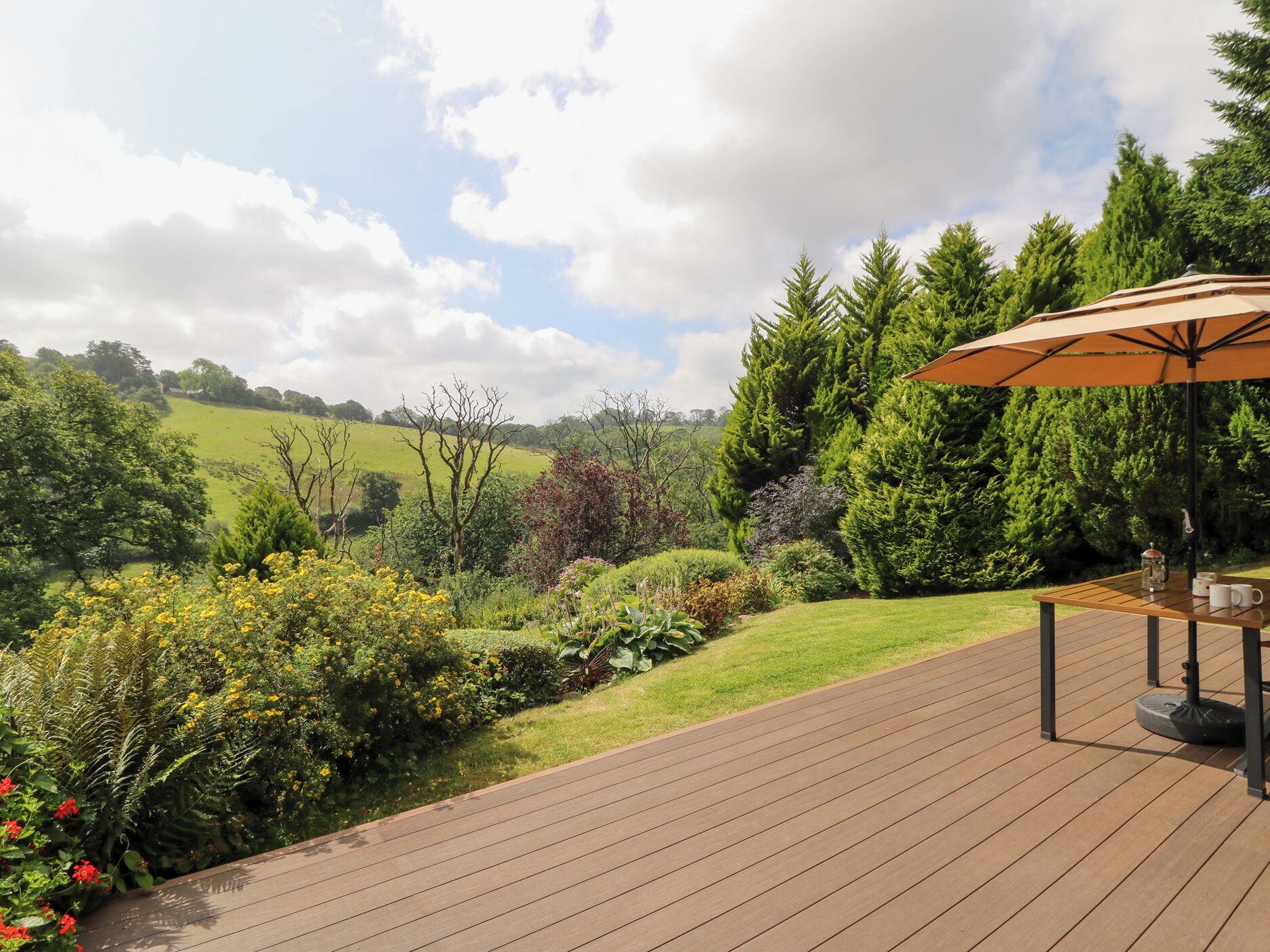 A wooden deck with a table and an umbrella, surrounded by lush greenery and a view of rolling hills.