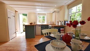 A cozy dining area with a wooden table set for tea, surrounded by a kitchen with a view of greenery. - Cwmbrandy Cottage (Fishguard)