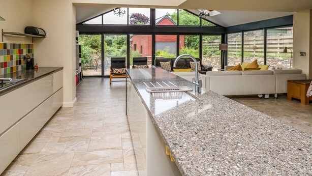 Una cocina moderna con una isla grande, un área de comedor con sofá y vistas a un jardín y una casa a través de ventanas amplias.