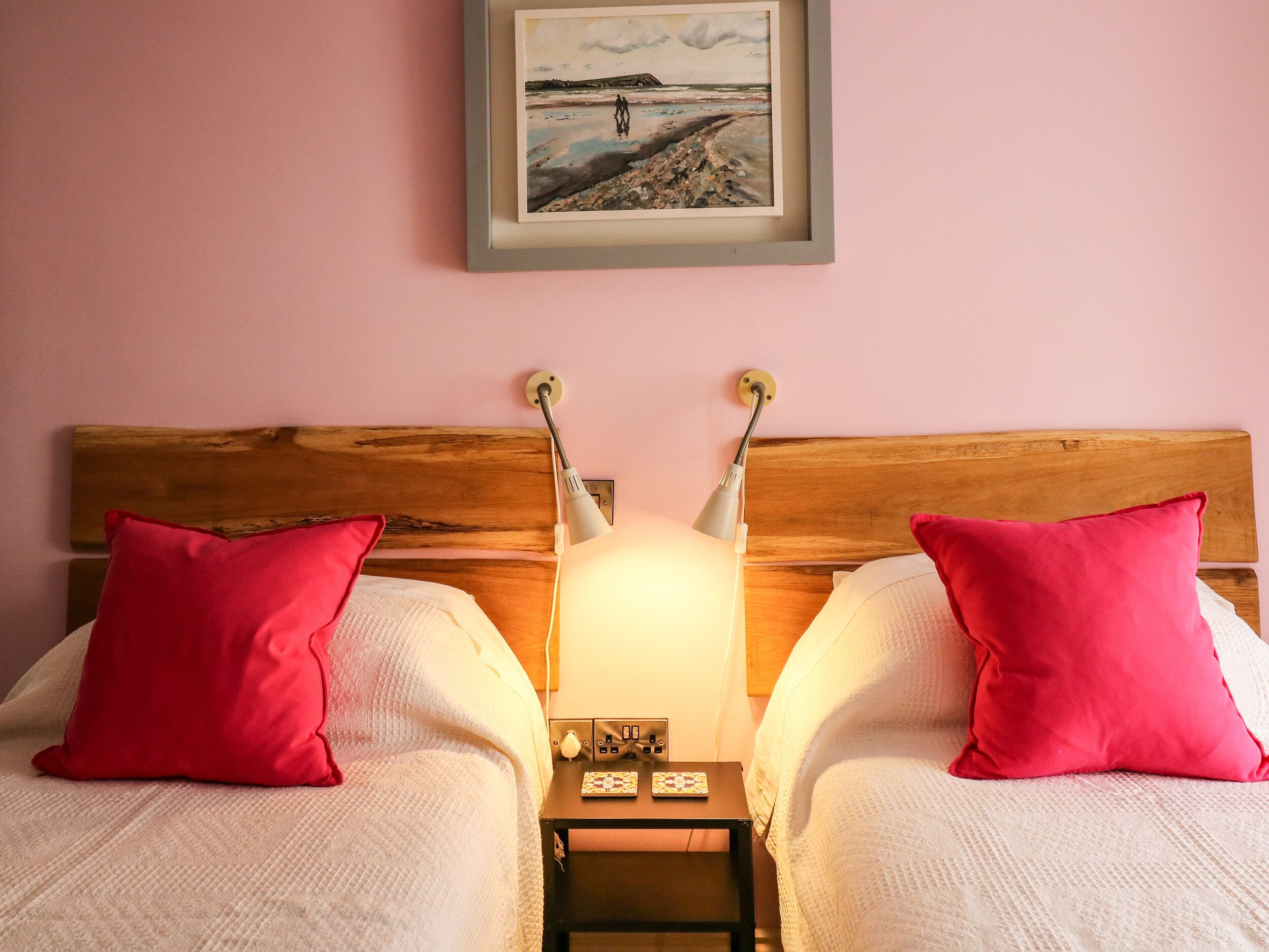 Un dormitorio con dos camas, cada una con un cojín rojo y una manta blanca. Entre las camas, un mesita de noche con una lámpara y un pequeño reloj. En la pared, sobre las camas, cuelga un cuadro enmarcado con una escena de playa.