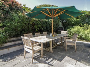 A wooden table with chairs and a green umbrella, surrounded by stone tiles and greenery.