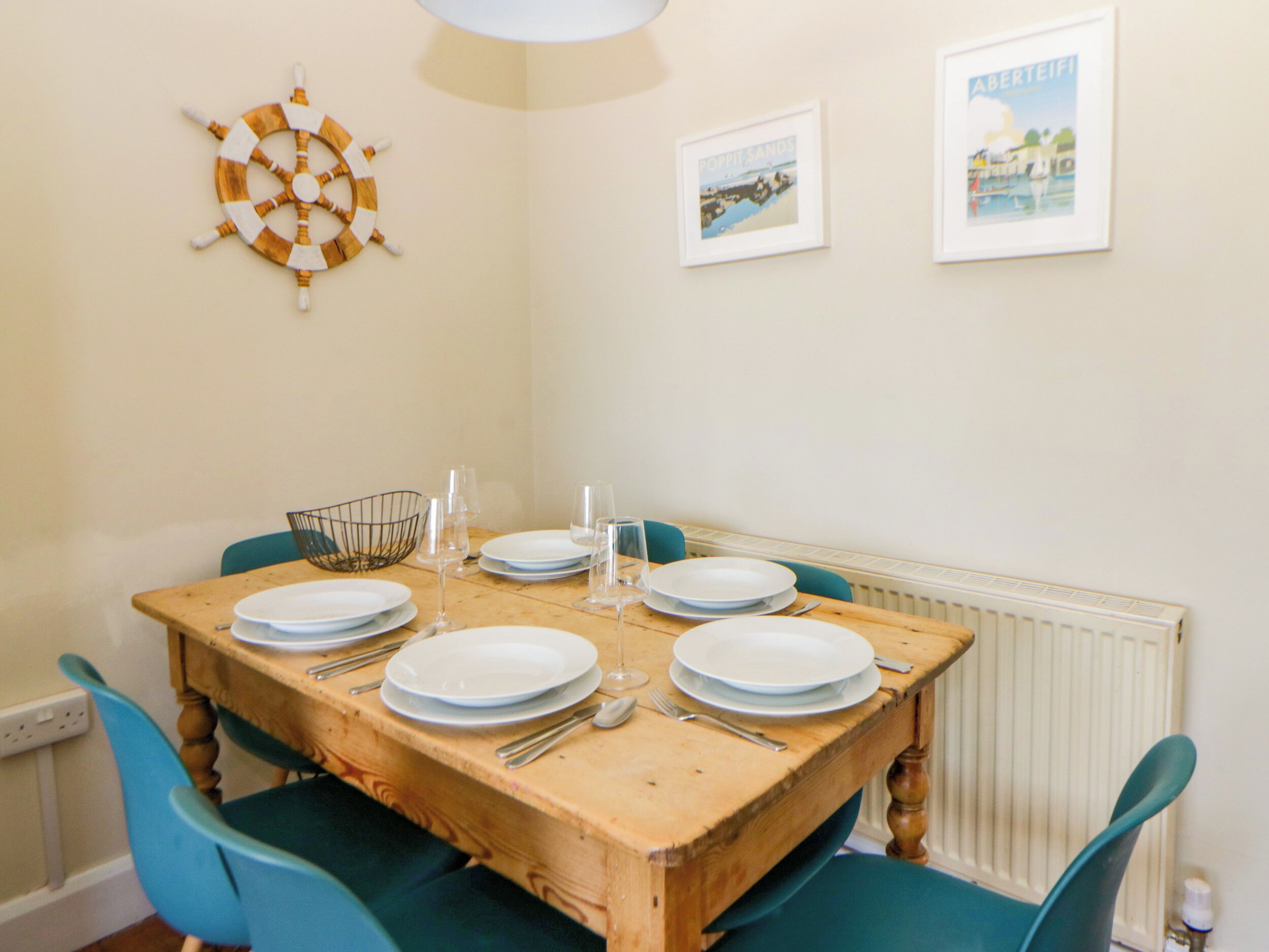 A dining table set with plates, glasses, and cutlery, surrounded by teal chairs, with a ship's wheel and framed pictures on the wall.