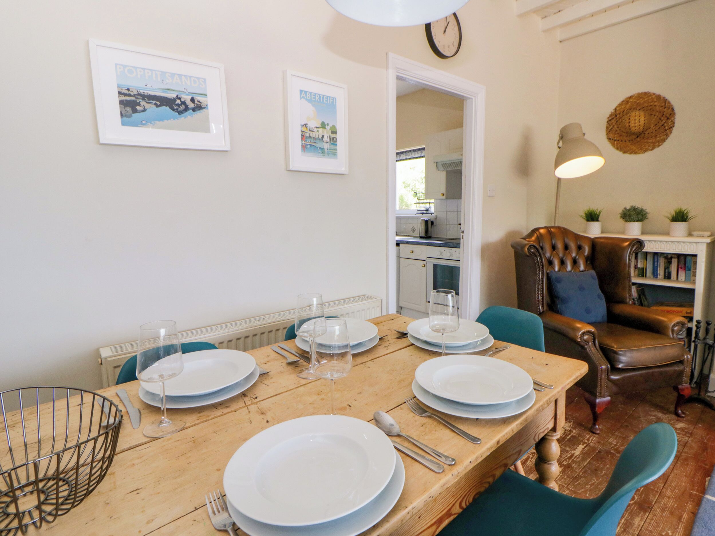Un coin repas avec une table en bois dressée pour quatre personnes, un panier en métal et une horloge au mur.